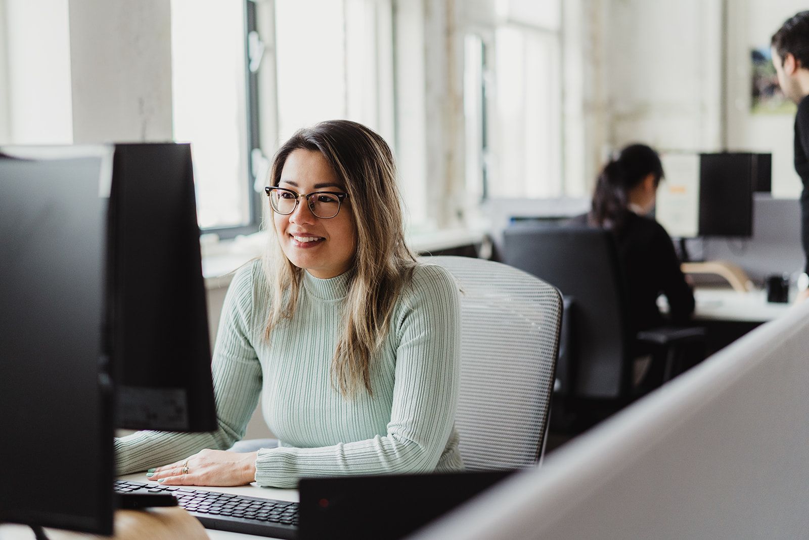  Een vrouw met een bril en een lichtgroene trui glimlacht terwijl ze aan een computer werkt in een lichte kantoorruimte, met een andere persoon op de achtergrond.